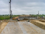 A construction site with workers engaged in building activity. Several large machines are visible, including cranes and excavators. The site appears expansive, with a partly constructed structure in the center. The ground is covered with sand and metal grates, and workers in safety vests are visible in the foreground. A green landscape with trees is visible in the background under a cloudy sky.