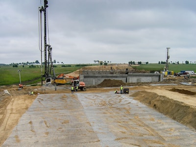 A construction site with workers engaged in building activity. Several large machines are visible, including cranes and excavators. The site appears expansive, with a partly constructed structure in the center. The ground is covered with sand and metal grates, and workers in safety vests are visible in the foreground. A green landscape with trees is visible in the background under a cloudy sky.