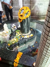 A stone Shiva lingam adorned with vibrant orange marigold garlands is placed in the center of a small pool surrounded by offerings including fruit and flowers. There are marble structures nearby, intricately decorated with similar offerings. A few people, partially visible, seem to be participating in or observing a ritual.