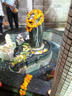A stone Shiva lingam adorned with vibrant orange marigold garlands is placed in the center of a small pool surrounded by offerings including fruit and flowers. There are marble structures nearby, intricately decorated with similar offerings. A few people, partially visible, seem to be participating in or observing a ritual.