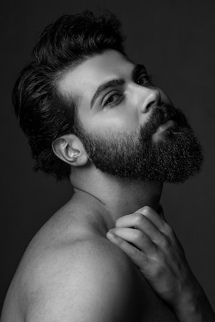 A black and white portrait of a bearded man with dark hair, tilting his head back slightly and looking towards the camera. The lighting highlights his facial features and the texture of his hair and beard. His hand gently rests on his neck, adding a sense of elegance to the composition.
