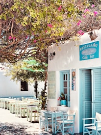 A charming outdoor seating area of a cafe with white wooden chairs and tables under a lush canopy of green and pink bougainvillea flowers. The cafe facade is white with light blue shutters and a sign that reads 'Restaurant, Cafe Bar'. There's a cozy, inviting atmosphere with natural light filtering through the foliage.