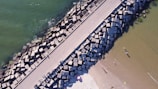 Aerial view of a coastal scene featuring a long concrete pier flanked by large, geometric wave breakers. The left side showcases green ocean water, while the right side has a sandy beach with people enjoying the shoreline. Some objects on the beach seem to include a bright pink umbrella and a surfboard or float.