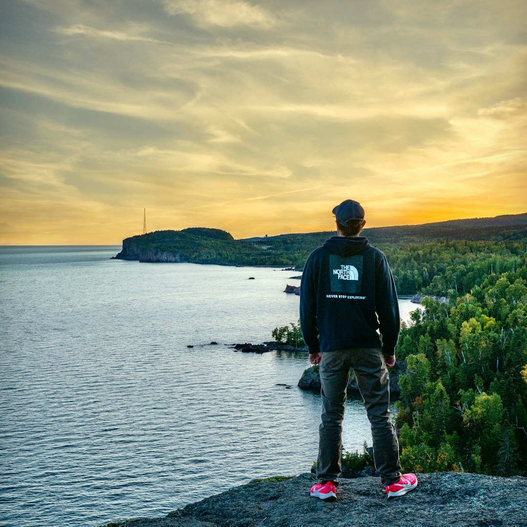 man in black jacket and blue denim jeans standing on rock near body of water during