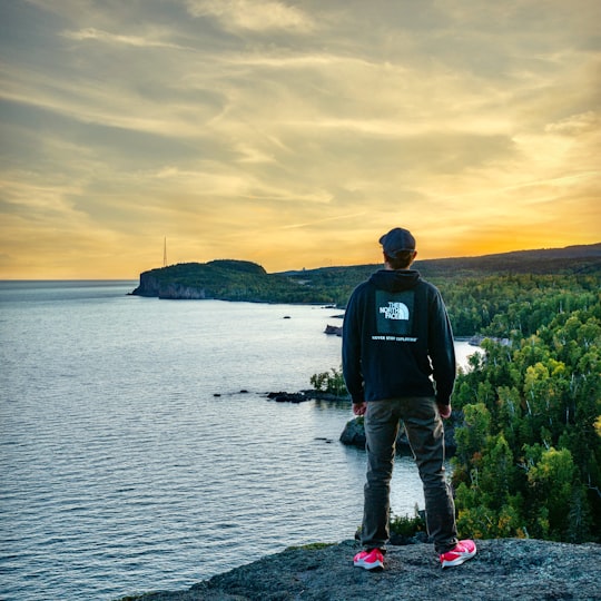 man in black jacket and blue denim jeans standing on rock near body of water during in Tettegouche State Park United States
