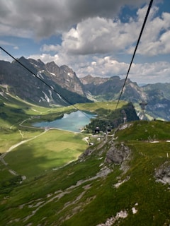A scenic mountainous landscape featuring a cable car system traversing over lush green hills with a stunning lake nestled in the valley below. Towering peaks surround the area, and the sky is partly cloudy, casting dynamic shadows over the terrain.