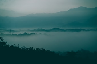 A serene landscape features layers of mist enveloping rolling hills, with the outline of mountains visible in the background. The atmosphere is calm and tranquil, drawing viewers into its mysterious ambiance.