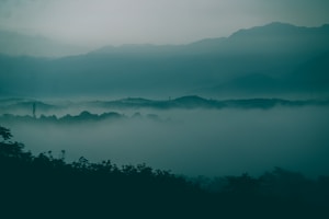 A serene landscape features layers of mist enveloping rolling hills, with the outline of mountains visible in the background. The atmosphere is calm and tranquil, drawing viewers into its mysterious ambiance.