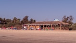 A beachside shack with a thatched roof is surrounded by palm trees under a clear blue sky. Sun loungers with red umbrellas are placed on the sandy beach in front of the shack.