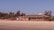 A beachside shack with a thatched roof is surrounded by palm trees under a clear blue sky. Sun loungers with red umbrellas are placed on the sandy beach in front of the shack.