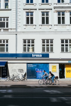 A street scene featuring a building with classical architectural elements, large windows, and a sign labeled 'Irma.' Bicycles are parked along the sidewalk, and two cyclists are passing by. A colorful advertisement is visible on the wall next to a discounted sales sign.