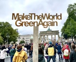 A large crowd of people gathered at a public demonstration, holding a large wooden sign that reads 'MakeTheWorldGreenAgain' in front of a historic landmark. Trees surround the area and the sky is overcast, adding a calm yet determined atmosphere.