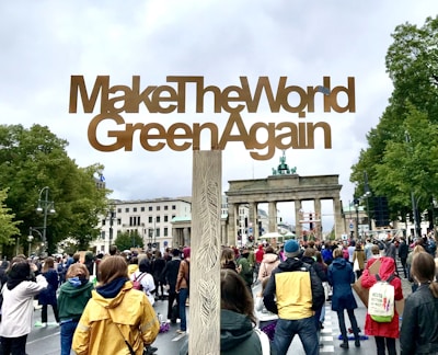 A large crowd of people gathered at a public demonstration, holding a large wooden sign that reads 'MakeTheWorldGreenAgain' in front of a historic landmark. Trees surround the area and the sky is overcast, adding a calm yet determined atmosphere.