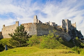 Ancient stone castle ruins stand prominently atop a grassy hill, surrounded by green shrubs and trees. The weathered walls and towers exhibit a sense of historical grandeur. The sky is blue with wispy white clouds, adding to the scene's expansive feeling.