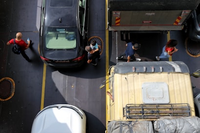 Aerial view of a parking area with several vehicles, including a black car, a white car, and a yellow truck parked closely together. Three people are walking near the vehicles, one holding a water bottle and the others moving in a different direction. The area is marked by yellow lines that separate the parking spaces.