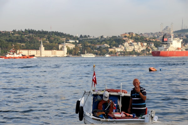 A small boat with a person standing aboard is in the foreground, floating on a large body of water. The person appears to be gesturing towards the camera, and there is a Turkish flag on the boat. In the background, a cityscape stretches across the shoreline with buildings, greenery, and a large cargo ship.