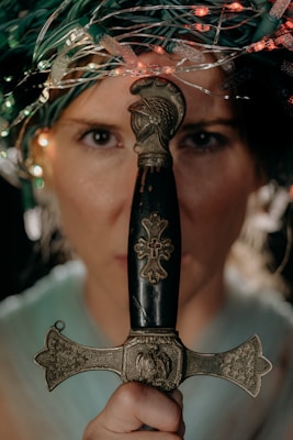 A person holds an ornate dagger vertically, with intricate details on the handle and crossguard. The person's face is partially obscured, and colorful lights or fibres frame their head, adding a mysterious ambiance.