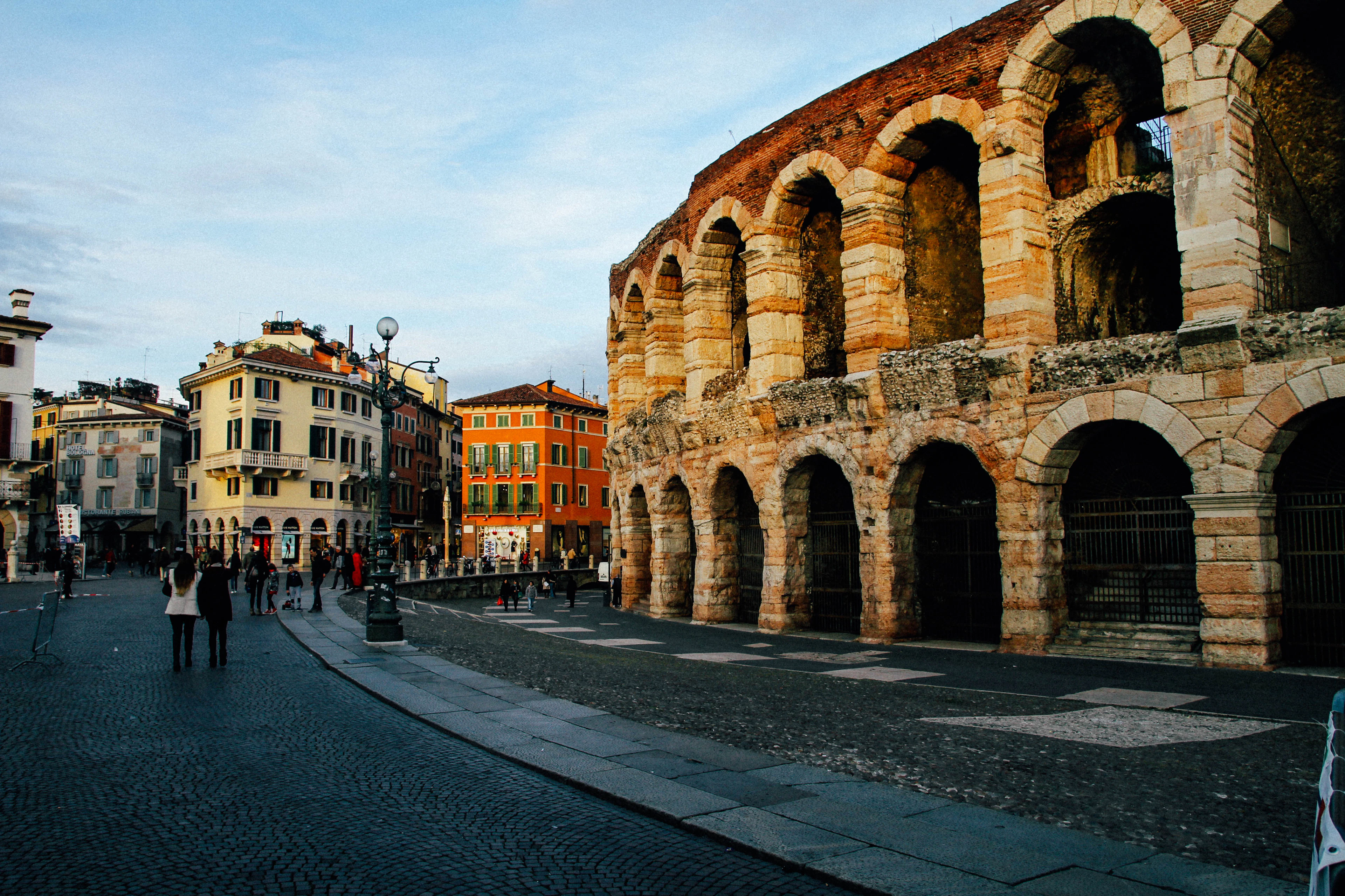 The stone arches of the Verona Arena glowing in evening light on Piazza Bra, with pastel buildings across the square