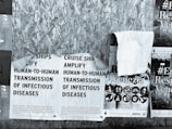 A series of overlapping posters attached to a wooden surface. The primary poster reads about the amplification of infectious disease transmission on cruise ships. Other posters feature graphic icons and the word 'Venezia,' along with partial images of warnings or advertisements. The scene is characterized by a textured wooden background.