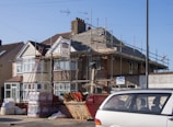 A two-story house undergoing roofing construction, with scaffolding surrounding the upper part of the building. Various construction materials, including wooden planks and boxes, are scattered around the site. A red skip and a parked car are visible in the foreground. The sky is clear and blue, indicating daytime.