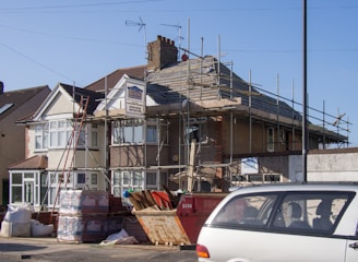 A two-story house undergoing roofing construction, with scaffolding surrounding the upper part of the building. Various construction materials, including wooden planks and boxes, are scattered around the site. A red skip and a parked car are visible in the foreground. The sky is clear and blue, indicating daytime.