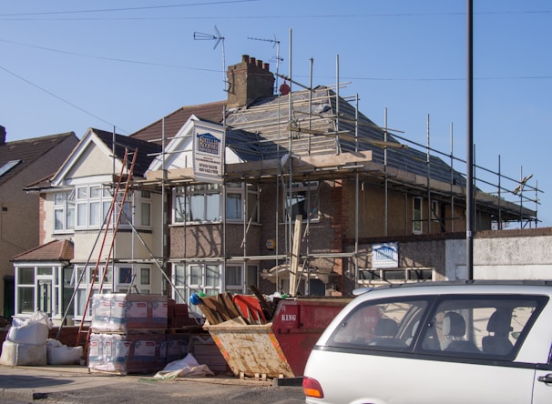 A two-story house undergoing roofing construction, with scaffolding surrounding the upper part of the building. Various construction materials, including wooden planks and boxes, are scattered around the site. A red skip and a parked car are visible in the foreground. The sky is clear and blue, indicating daytime.