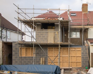A residential building under construction with visible scaffolding surrounding the front. The structure features a mix of bare brickwork and wooden panels. Windows are installed on the upper level, and the roof is partially finished with red tiles. Construction materials such as wooden planks and blue tarps are scattered in the foreground.