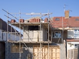 Scaffolding surrounds a house under construction or renovation, featuring brickwork with unfinished sections near the roof. A standard two-story building shows the first-floor structure partially completed. Roofing tiles are piled on scaffolding positioned above the front of the house.