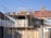 Scaffolding surrounds a house under construction or renovation, featuring brickwork with unfinished sections near the roof. A standard two-story building shows the first-floor structure partially completed. Roofing tiles are piled on scaffolding positioned above the front of the house.