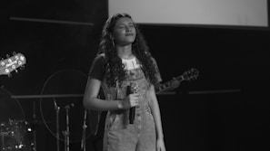 A young person with long curly hair stands on a dimly lit stage holding a microphone, eyes closed, possibly singing or emoting intensely. The setting appears to be a music performance as there are instruments visible in the background, including a guitar and part of a drum set.