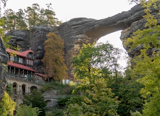 A large natural rock arch formation towers over a lush, green forest. Beside the arch, a rustic building with a red roof nestles against the rock face, while mature trees surround the scene, indicating a serene and secluded setting.