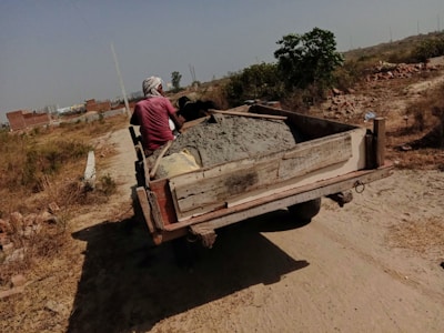 A person is driving a cart filled with cement along a dirt path. The surrounding area is rural with sparse vegetation, and a few unfinished brick structures can be seen in the background.