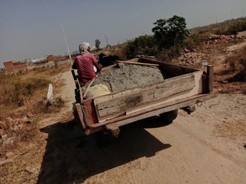 A person is driving a cart filled with cement along a dirt path. The surrounding area is rural with sparse vegetation, and a few unfinished brick structures can be seen in the background.