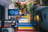 An eclectic stairway leads up to a cozy cafe and bistro. Bright neon signs advertise a hotel, nargile cafe, and water pipes. The vibrant atmosphere is enhanced by colorful decorations and potted plants, creating a welcoming ambiance. A couple of patrons are seated at a table, adding a sense of leisurely enjoyment to the scene.