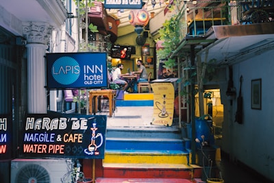 An eclectic stairway leads up to a cozy cafe and bistro. Bright neon signs advertise a hotel, nargile cafe, and water pipes. The vibrant atmosphere is enhanced by colorful decorations and potted plants, creating a welcoming ambiance. A couple of patrons are seated at a table, adding a sense of leisurely enjoyment to the scene.