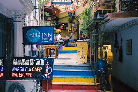 An eclectic stairway leads up to a cozy cafe and bistro. Bright neon signs advertise a hotel, nargile cafe, and water pipes. The vibrant atmosphere is enhanced by colorful decorations and potted plants, creating a welcoming ambiance. A couple of patrons are seated at a table, adding a sense of leisurely enjoyment to the scene.