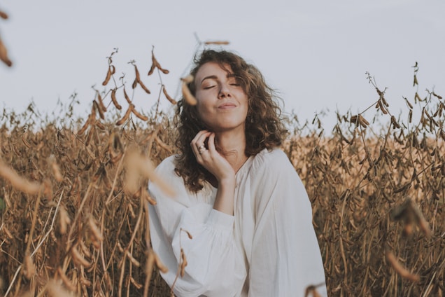 A person with curly hair is standing in a field of dried plants, dressed in a white garment. Eyes are closed, conveying a sense of peace and relaxation.
