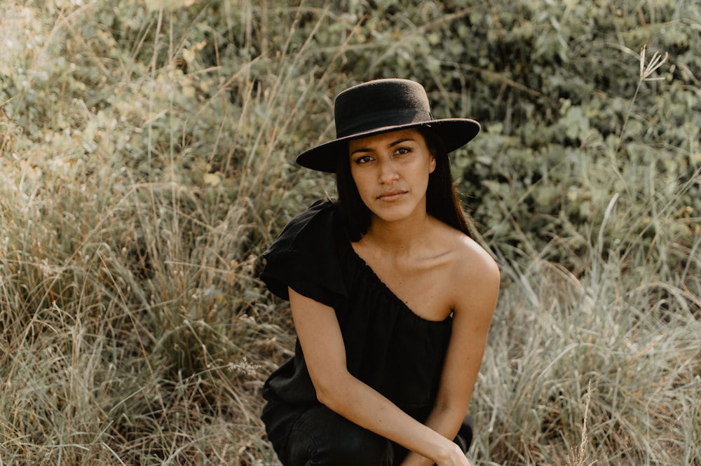 woman in black off shoulder dress wearing black hat sitting on brown grass field during daytime
