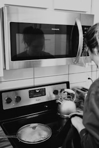 A woman stands at a modern kitchen stove, wearing casual attire. Reflected in the microwave above, her face is faintly visible. A stainless steel kettle is boiling on the stove next to a covered pot. The kitchen appliances include a Frigidaire microwave and a Whirlpool stove, with a digital display showing '160'.