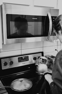 A woman stands at a modern kitchen stove, wearing casual attire. Reflected in the microwave above, her face is faintly visible. A stainless steel kettle is boiling on the stove next to a covered pot. The kitchen appliances include a Frigidaire microwave and a Whirlpool stove, with a digital display showing '160'.
