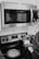 A woman stands at a modern kitchen stove, wearing casual attire. Reflected in the microwave above, her face is faintly visible. A stainless steel kettle is boiling on the stove next to a covered pot. The kitchen appliances include a Frigidaire microwave and a Whirlpool stove, with a digital display showing '160'.