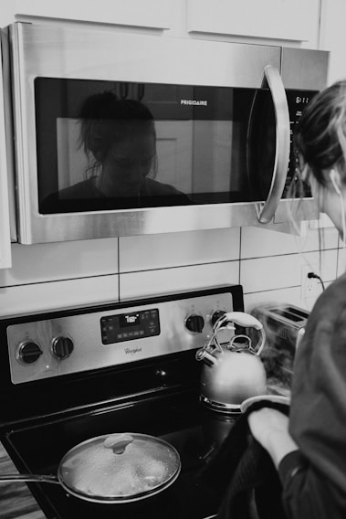A woman stands at a modern kitchen stove, wearing casual attire. Reflected in the microwave above, her face is faintly visible. A stainless steel kettle is boiling on the stove next to a covered pot. The kitchen appliances include a Frigidaire microwave and a Whirlpool stove, with a digital display showing '160'.