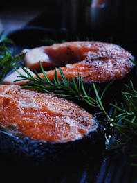 Two raw salmon steaks are placed on a black cooking surface, garnished with fresh green rosemary sprigs. The salmon appears fresh with a vibrant orange-pink color and scattered specks of seasoning, likely salt and pepper, on top.