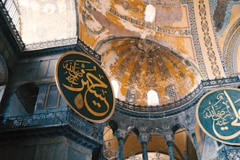 The image features an intricately decorated interior of a historical building, showcasing large circular plaques with Arabic calligraphy. The architecture includes high domes with ornate designs and faded frescoes, illuminated by natural light from windows.