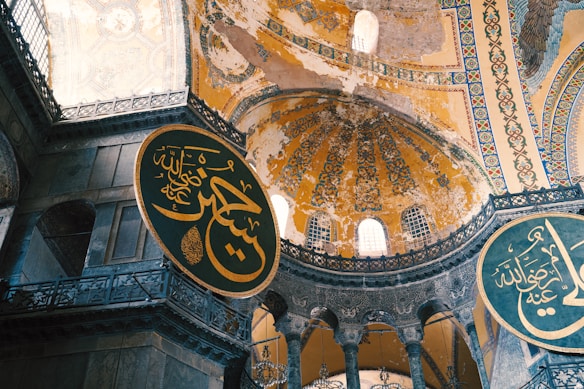 The image features an intricately decorated interior of a historical building, showcasing large circular plaques with Arabic calligraphy. The architecture includes high domes with ornate designs and faded frescoes, illuminated by natural light from windows.