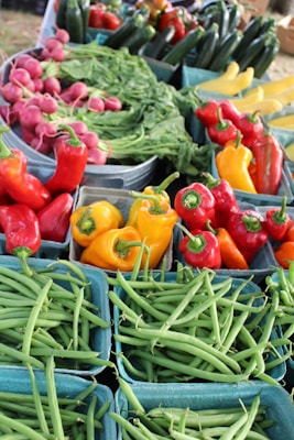 A vibrant assortment of fresh vegetables arranged in baskets. There are red and yellow bell peppers, green beans, radishes, leafy greens, and cucumbers.