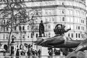 A black and white photo focuses on a statue of a mythological figure holding a fish, with a large, historic building in the background. The building has numerous windows illuminated, suggesting it is evening or night. There are trees and people walking on the street, giving the scene a bustling urban vibe.