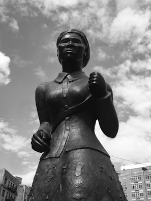 A black and white photograph of a bronze statue depicting a woman in a historical outfit. The statue is positioned against a partly cloudy sky, with buildings visible in the background. The woman appears determined, with a clenched fist and a focused expression.