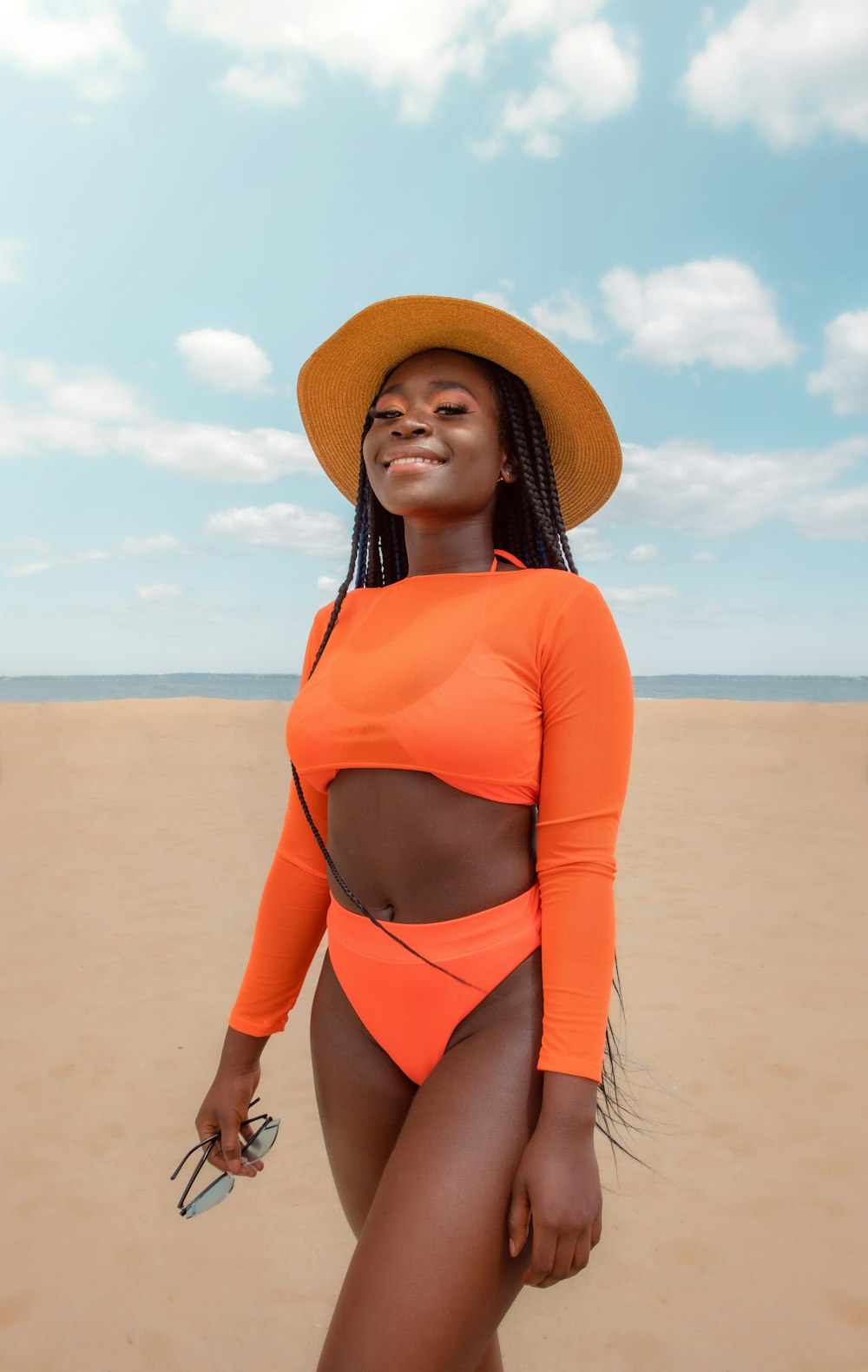オレンジ色の長袖シャツと茶色の日よけ帽をかぶった女性が、昼間、茶色の砂浜に立っている