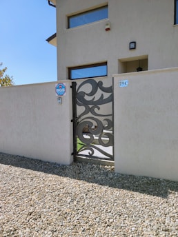 A modern house facade with a decorative metal gate set within a beige wall. The gate has intricate swirling patterns and is complemented by small gravel stones in the foreground. There are minimalistic rectangular windows, and a sign with 'BGS' nearby. The building number '25C' is visible.